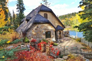Une maison en pierre avec un toit à forte pente, une clôture en bois et un étang en arrière-plan.