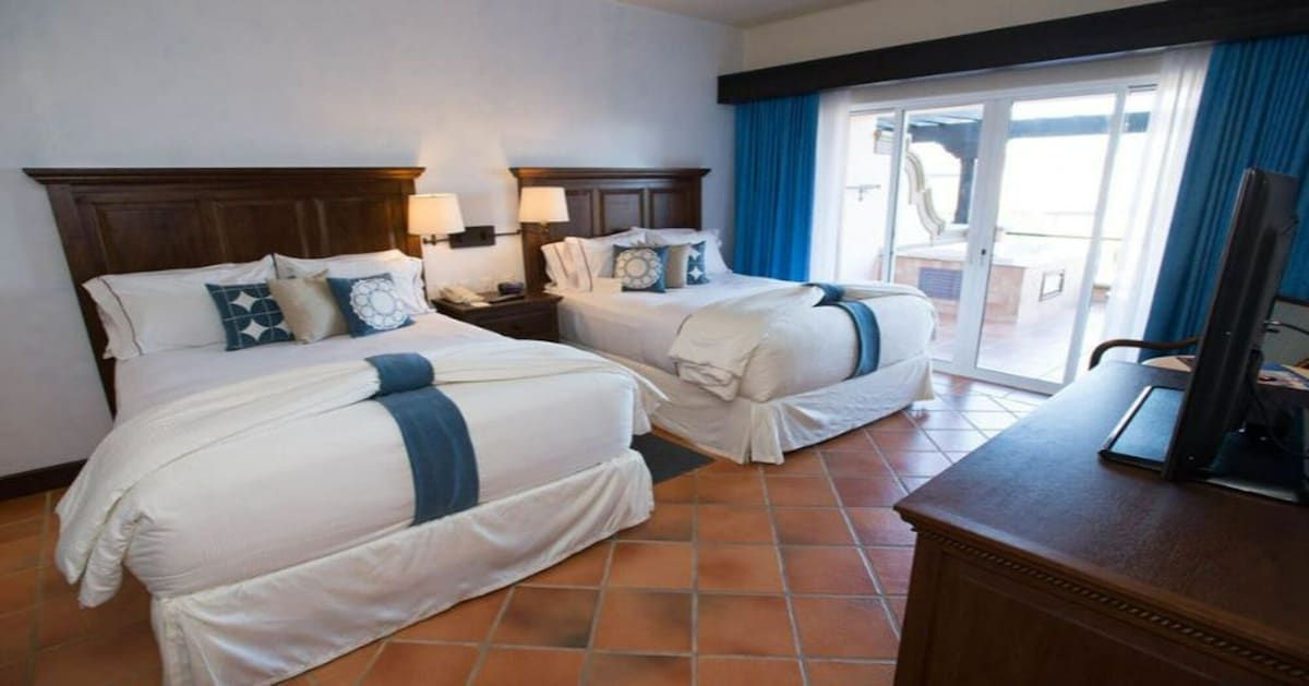 A hotel room with two beds, a desk, and a view of the outside.