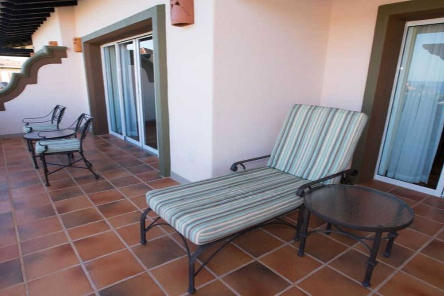 A covered patio area with a tiled floor, a striped lounge chair, a small round table, and a metal staircase.