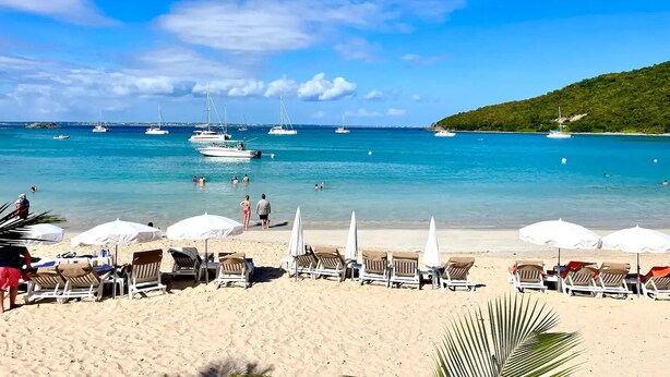 A beach with sun loungers and umbrellas, boats in the water, and a hilly landscape in the background.