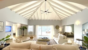 A spacious living room with a high ceiling, a ceiling fan, a large sofa set, a coffee table, and a television displaying 'ETFLIX'.