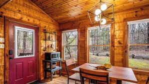 A wooden cabin interior with a red door, a dining table, chairs, a microwave, and shelves with various items.