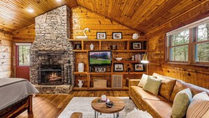 A log cabin interior with a stone fireplace, wooden walls, a sofa, and a television.