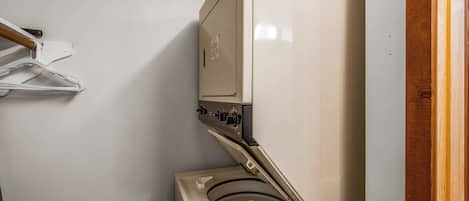 Una lavadora blanca y compacta con panel de control, una puerta de madera y un tendedero fijado a la pared.