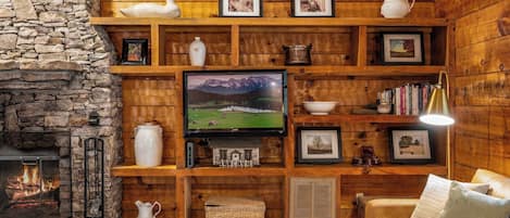 A cozy cabin interior with a stone fireplace, wooden shelves with various items, a television, and a comfortable seating area.