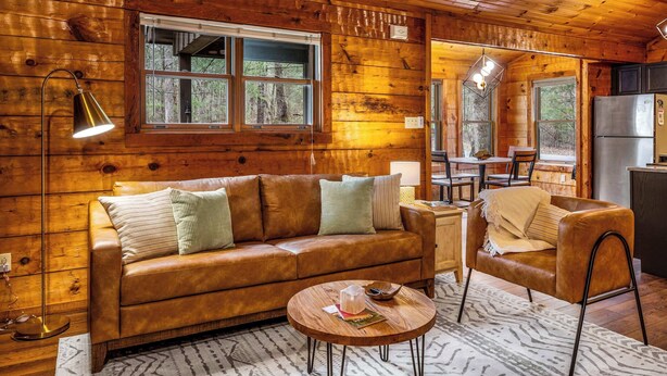 A wooden cabin interior with a leather sofa, a round coffee table, a dining table, and a kitchen area.
