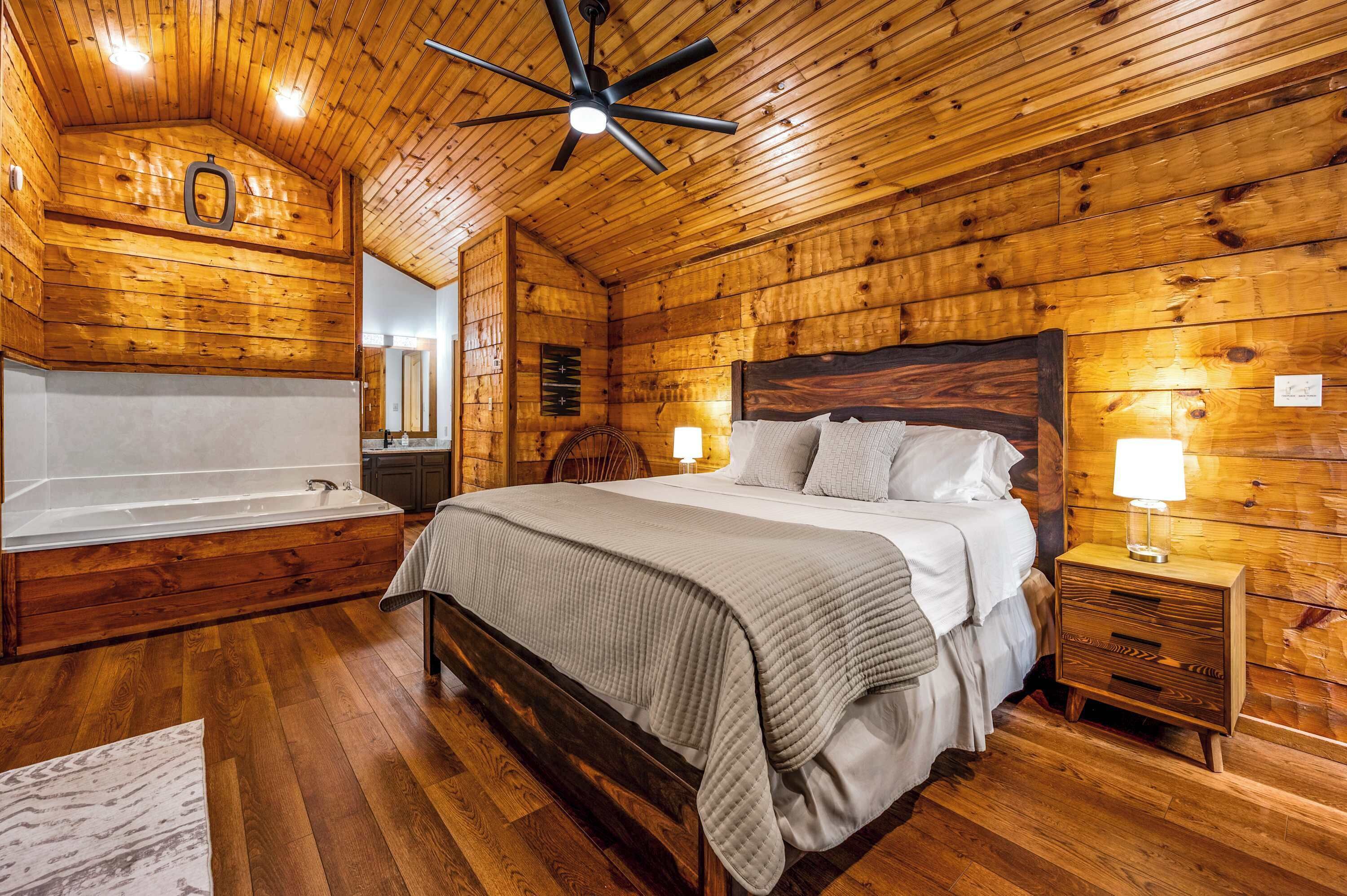 A wooden cabin bedroom with a large bed, a wooden headboard, a nightstand, a bedside lamp, a wooden ceiling fan, and a wooden bathtub.