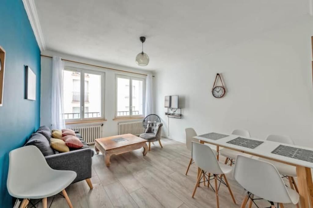 Una sala moderna con sofá, mesa de centro y mesa de comedor con sillas. Hay ventanas amplias, un radiador y un reloj fijado a la pared.