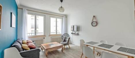 Una sala moderna con sofá, mesa de centro y mesa de comedor con sillas. Hay ventanas amplias, un radiador y un reloj fijado a la pared.