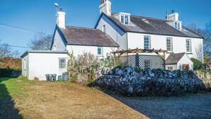 A white two-story house with a sloped roof, a small attached garage, and a garden area with a wooden fence. - Milan Cottage - 2 Bedroom Cottage - Port Eynon (Swansea)