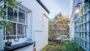 A narrow courtyard with a stone wall, a white building with a window, and a metal table and chair set. - Milan Cottage - 2 Bedroom Cottage - Port Eynon (Swansea)