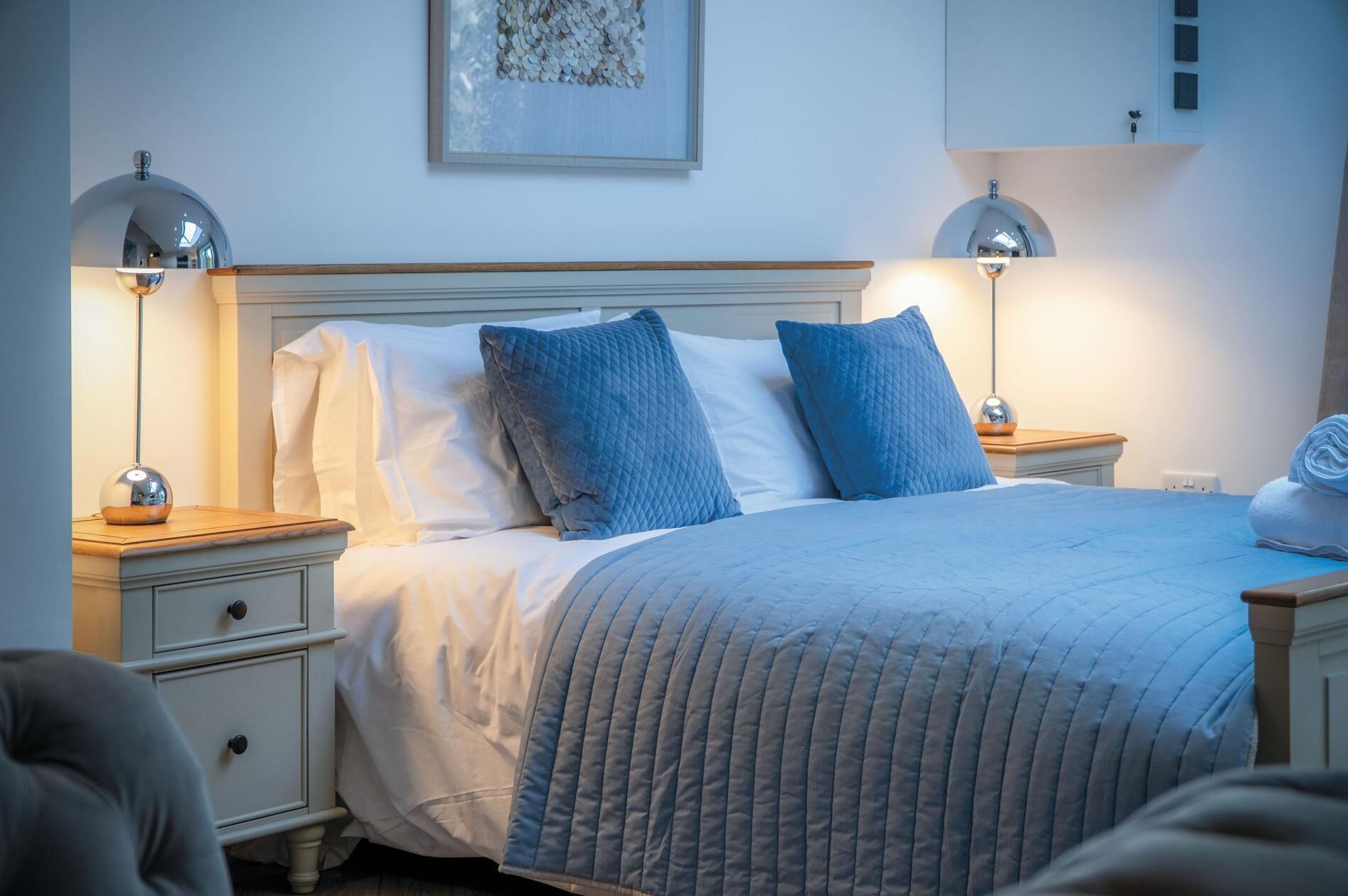 Un lit bien fait, recouvert d’une couverture bleue matelassée, avec deux oreillers bleus assortis et une couette blanche. On y trouve deux tables de chevet avec des lampes, une table de nuit en bois et une étagère fixée au mur.