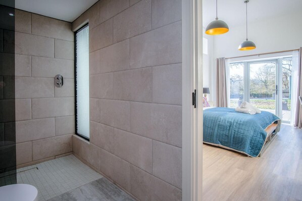 Un baño moderno con ducha, inodoro y lavamanos. El baño tiene un ventanal grande y una puerta que da a un dormitorio. El dormitorio tiene una cama, un ventanal y vistas al exterior.