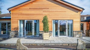 A modern wooden house with a gabled roof, large glass doors, and a central planter with a small tree.