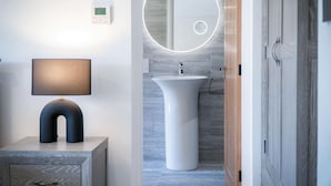 A modern bathroom with a round mirror, a white pedestal sink, and a wall-mounted thermostat. - Suite 10 - Sleeping Giant Hotel - Pen Y Cae Inn (Swansea Valley)