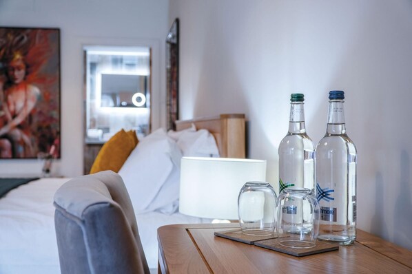 A hotel room with a bed, a wooden side table with bottles, a lamp, and a painting on the wall.