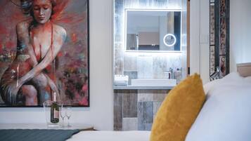 A hotel room with a large painting, a bed with a yellow pillow, a bedside table with a bottle and glasses, and a bathroom visible through a glass door.