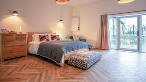 A modern bedroom with a herringbone wooden floor, a large bed with a blue quilt, a wooden dresser, and a view of the outdoors through a glass door. - Suite 4 - Sleeping Giant Hotel - Pen Y Cae Inn (Swansea Valley)