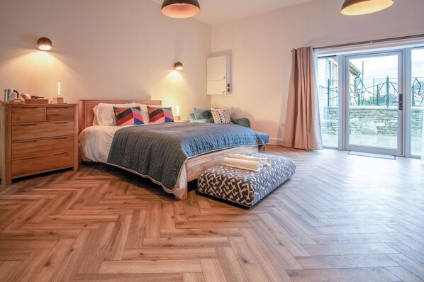 A modern bedroom with a herringbone wooden floor, a large bed with a blue quilt, a wooden dresser, and a view of the outdoors through a glass door.