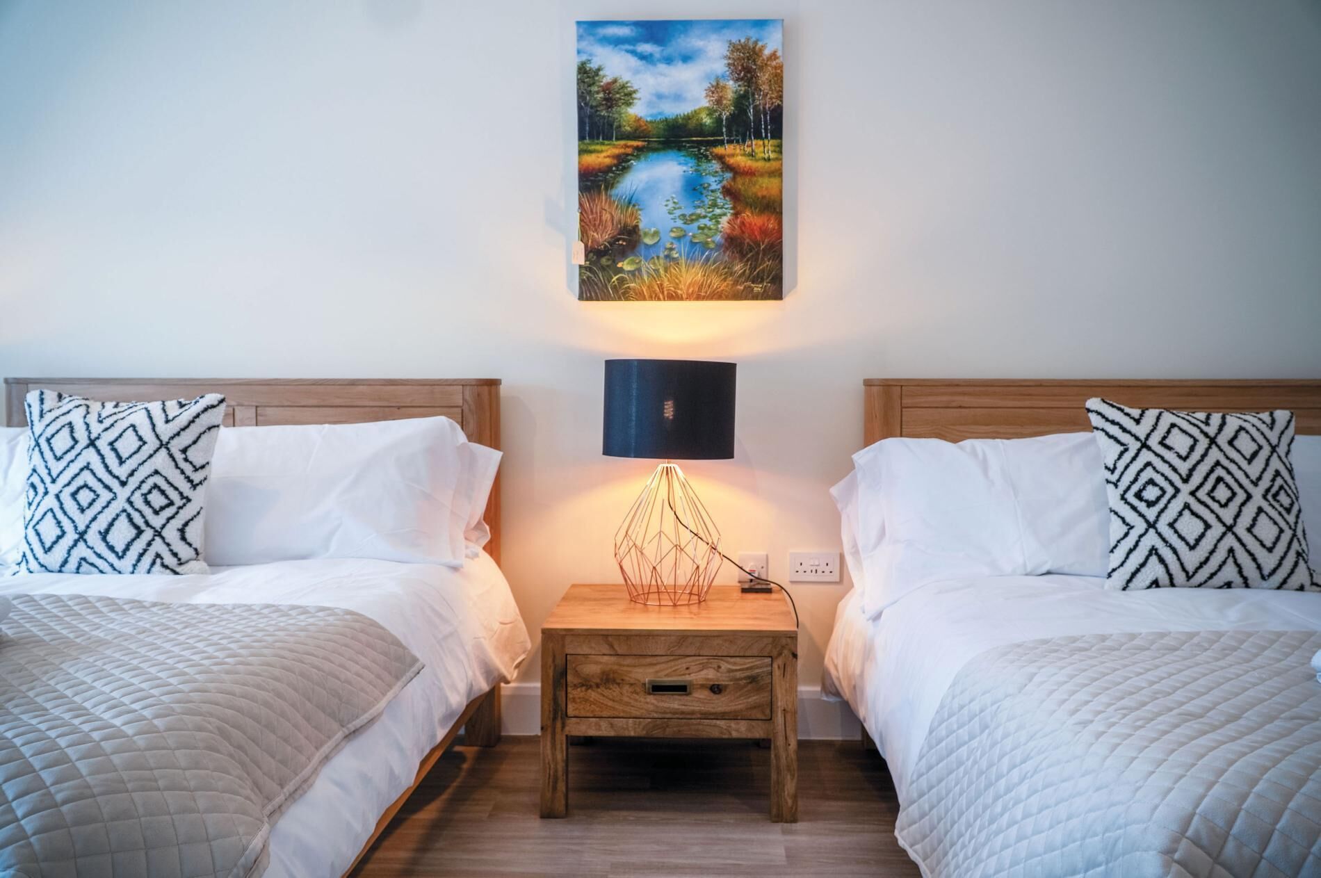 A hotel room with two beds, a nightstand with a lamp, and a painting of a landscape on the wall.