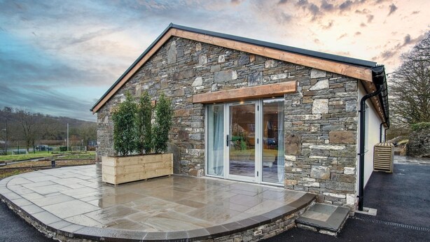 Un bâtiment en pierre avec un toit en bois, une porte en verre et un patio pavé agrémenté d’un jardinier en bois.