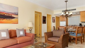 A living room with a sofa, armchair, and coffee table, a dining area with chairs and a table, and a kitchen with cabinets and a chandelier.