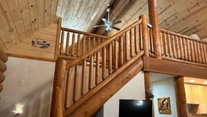 Un escalier en bois, un ventilateur de plafond, un téléviseur à écran plat fixé au mur et, au fond, un coin cuisine.