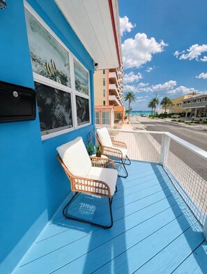 A balcony with two wicker chairs, a blue floor, and a view of a beach and buildings.