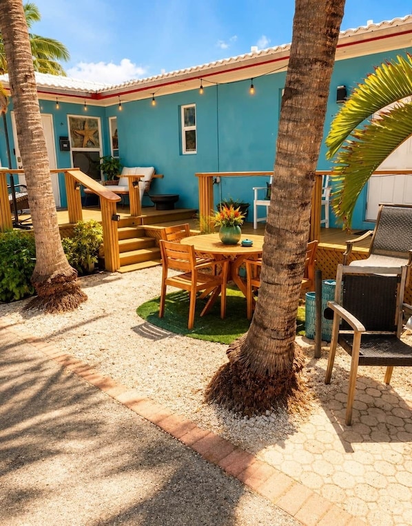 Eine Terrasse mit Holztisch und -stühlen, eine kleine Treppe und ein blaues Gebäude im Hintergrund.