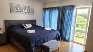 Ein Schlafzimmer mit einem großen Bett, einem Nachttisch, einer Bank, einem Gemälde und Blick auf Grün durch das Fenster.