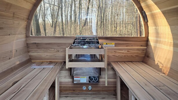 A wooden cabin with a central stove, benches, and a window overlooking a forest. - 14 Min to Lake Michigan: 3BR, 5 Beds, 30 Acres, Hot Tub, Sauna, Pets & Fire Pit (Covert)