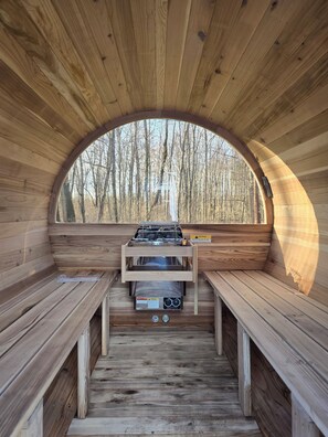 Een houten hut met een centraal geplaatste kachel, banken en een raam met uitzicht op een bos.