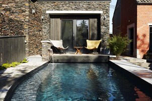 Una piscina con dos sillas de descanso y una mesita, rodeada de muros de piedra y una terraza de madera.
