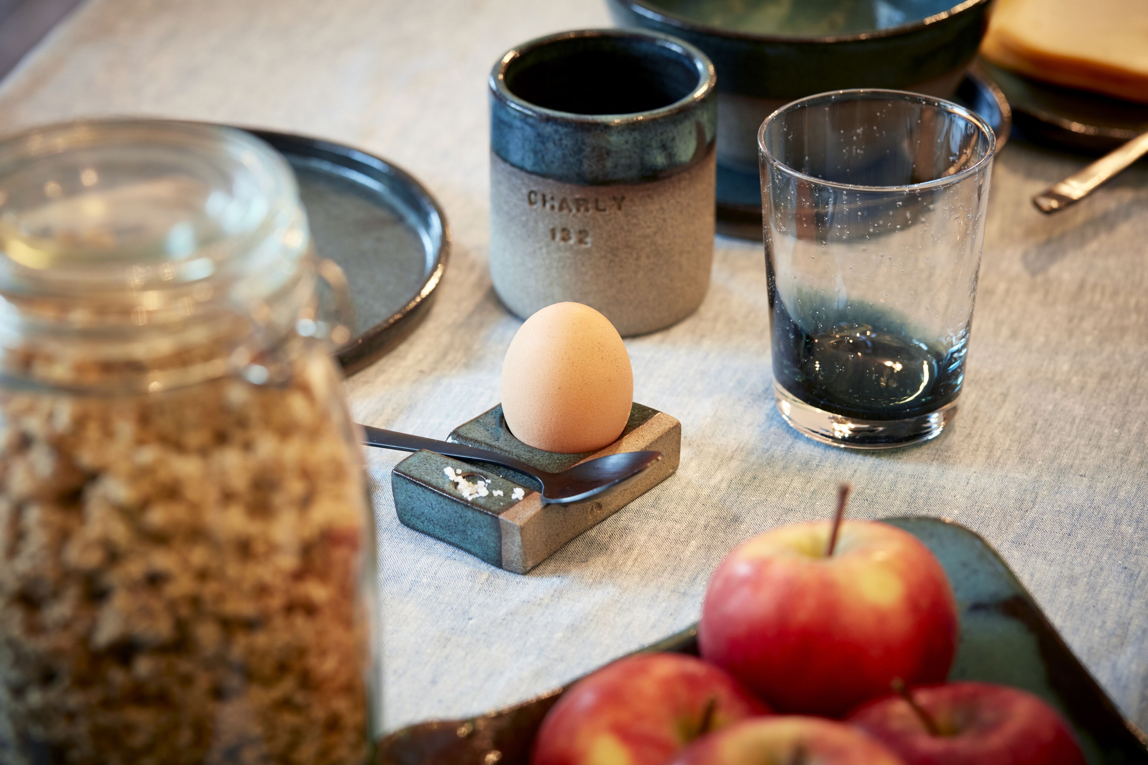 Una mesa con una taza de cerámica, un vaso, una cuchara, un huevo duro y manzanas.