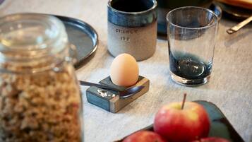 Una mesa con una taza de cerámica, un vaso, una cuchara, un huevo duro y manzanas.
