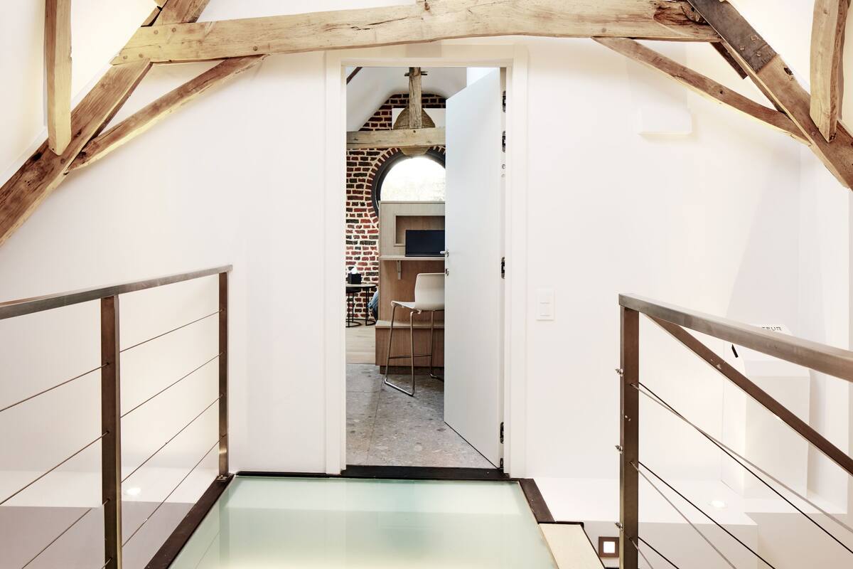 A modern interior with a glass floor leading to a lower level, wooden beams on the ceiling, and a brick wall in the background.