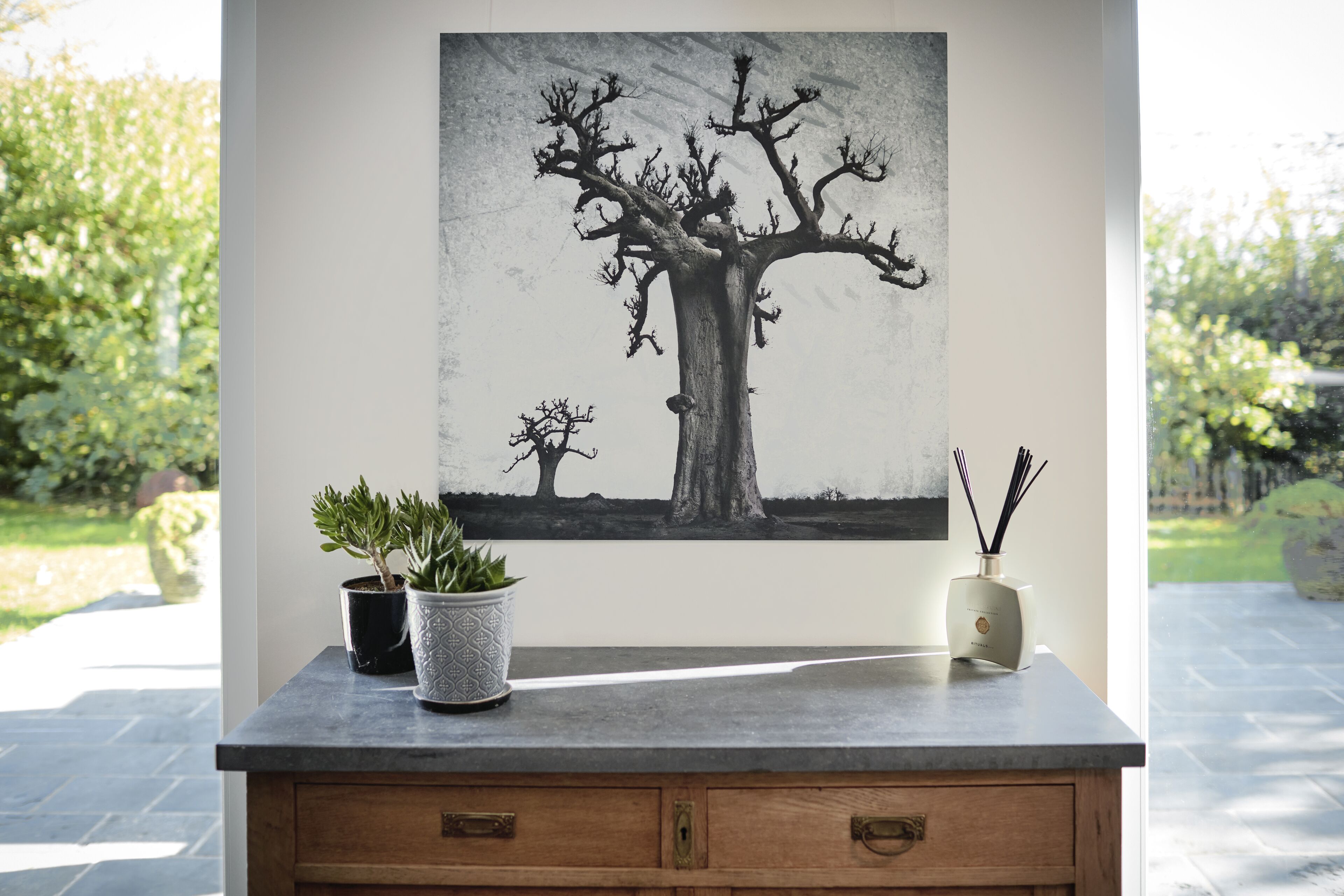 A room with a large painting of a tree on the wall, a wooden cabinet with drawers, and two potted plants on a counter.