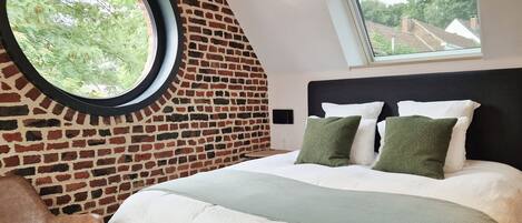 A bedroom with a brick wall, a round window, a bed with white and green bedding, and a view of trees and houses through the window.