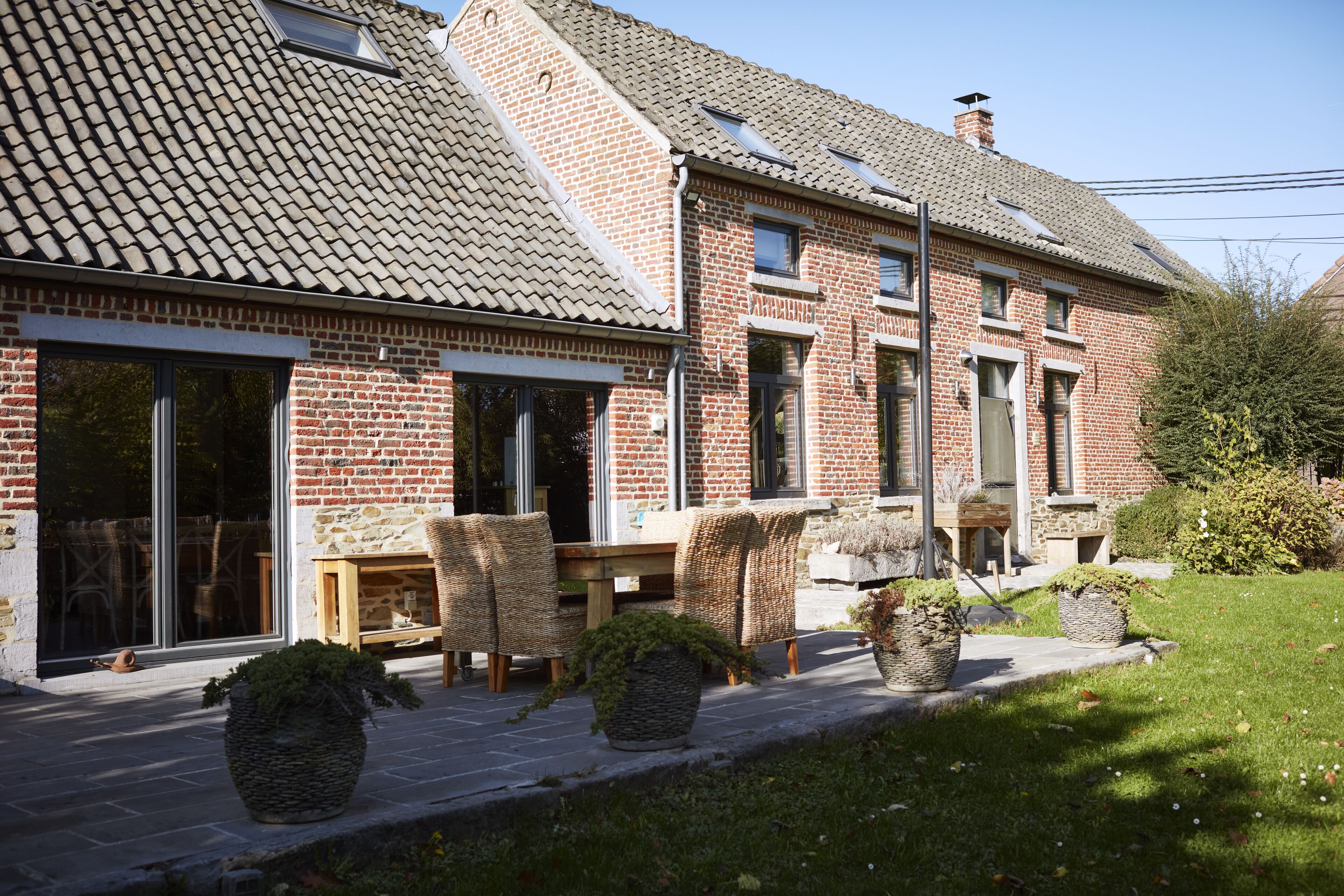A brick house with a tiled roof, large windows, and outdoor seating area with wicker chairs and potted plants.
