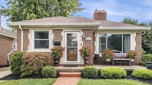 Una casa a un piano in mattoni con un portico, una cassetta delle lettere e un piccolo giardino con arbusti e una panchina.