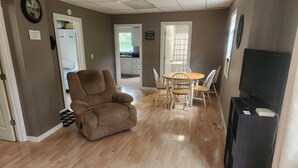 Un salon avec un fauteuil inclinable marron, une table à manger en bois avec quatre chaises, un téléviseur posé sur un meuble et une cuisine visible à travers une porte ouverte.