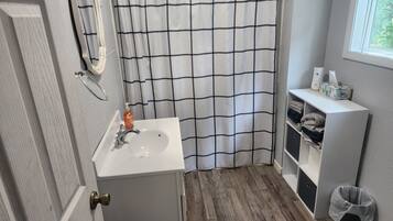 A bathroom with a white sink, a mirror, a shower curtain with a black grid pattern, a wooden floor, and a window with blinds.