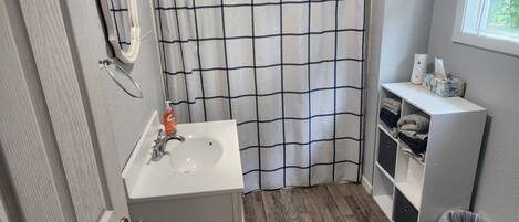 A bathroom with a white sink, a mirror, a shower curtain with a black grid pattern, a wooden floor, and a window with blinds.