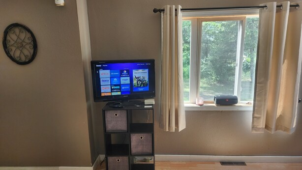 Un salon avec un téléviseur à écran plat posé sur un meuble, une fenêtre avec des rideaux et un objet décoratif fixé au mur.