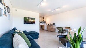 Ein modernes Wohnzimmer mit einem dunkelblauen Sofa, einem Couchtisch und einer großen Pflanze. Im Hintergrund ist eine Küche mit Essbereich zu sehen.