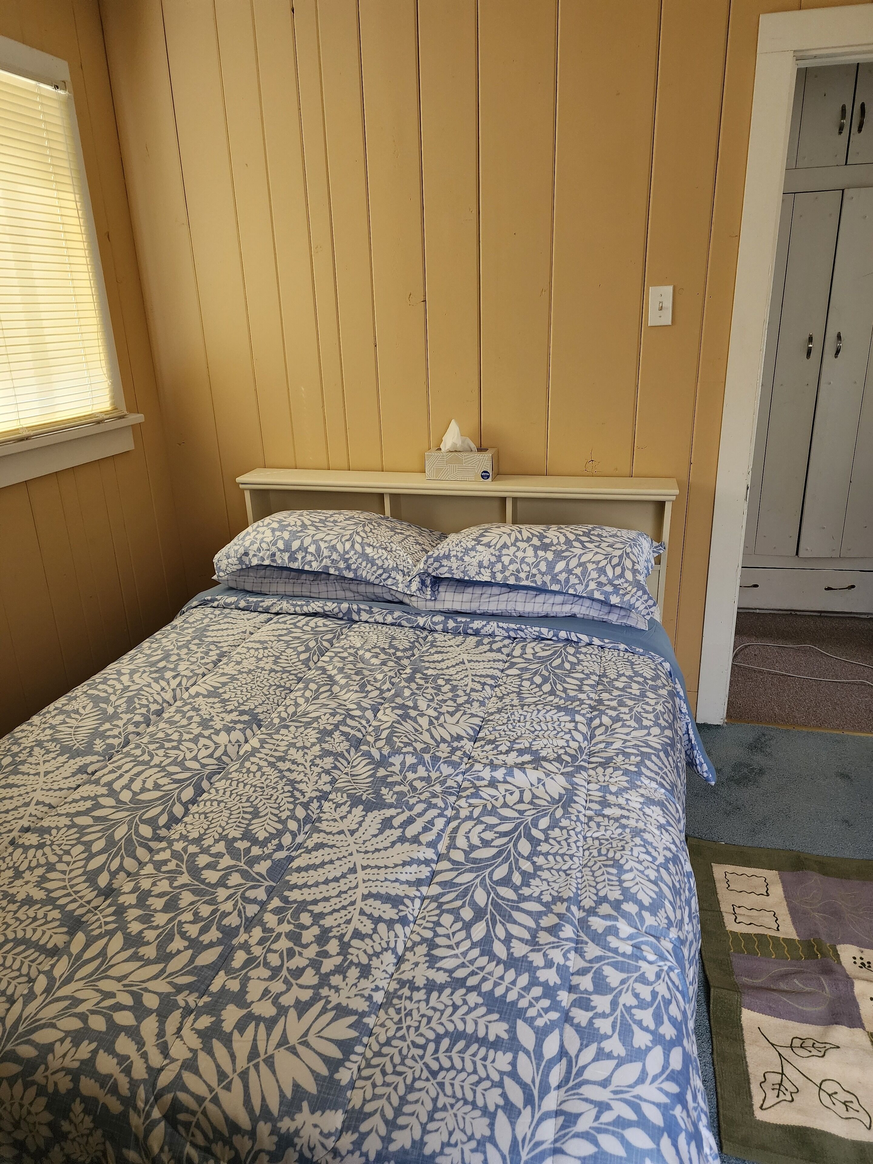 Ein Schlafzimmer mit Bett, Fenster mit Jalousien, einer Taschentuchbox auf einem Regal und einem Kleiderschrank.