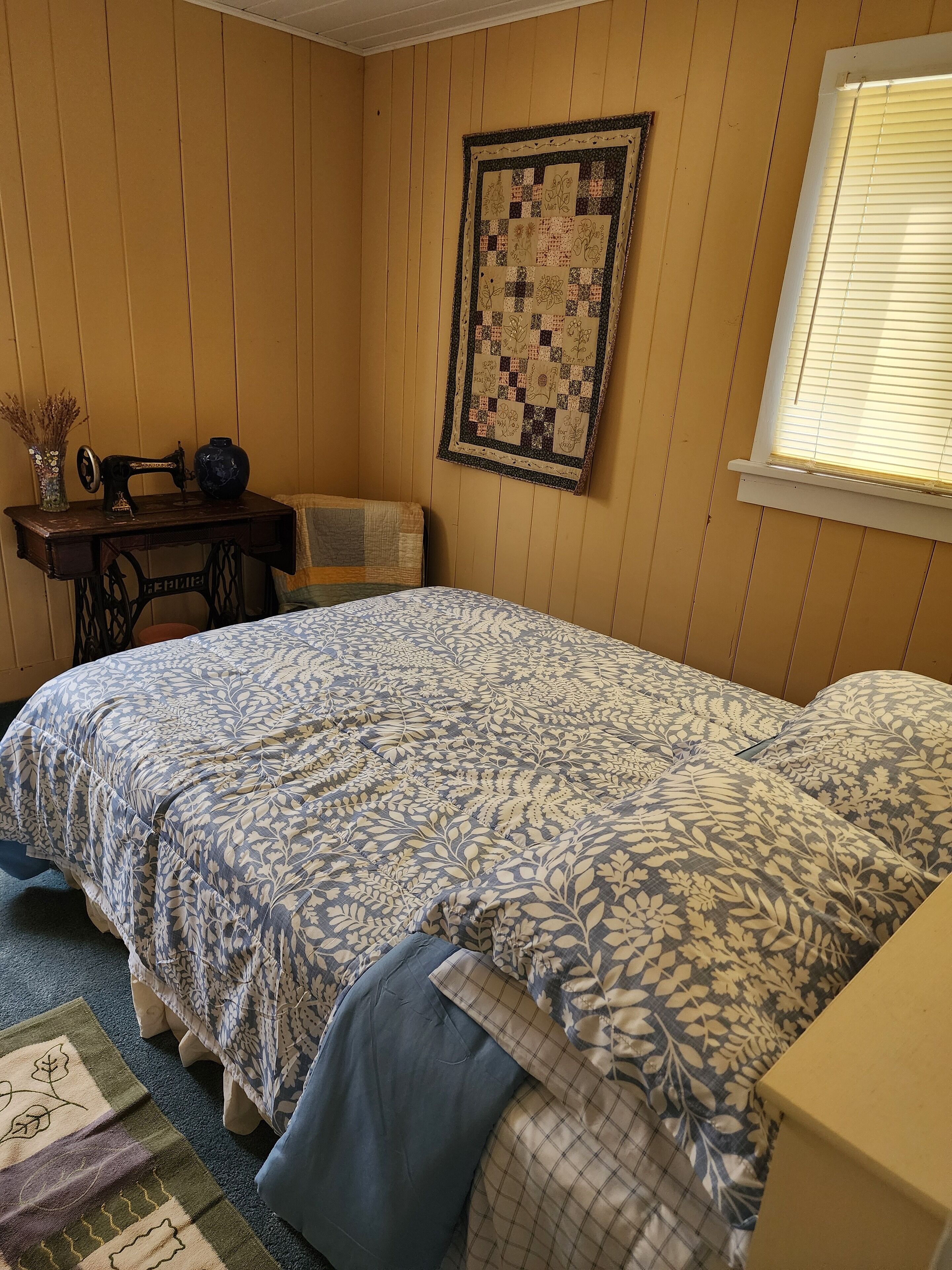 Une chambre à coucher avec un lit, une couverture, une machine à coudre et une fenêtre.