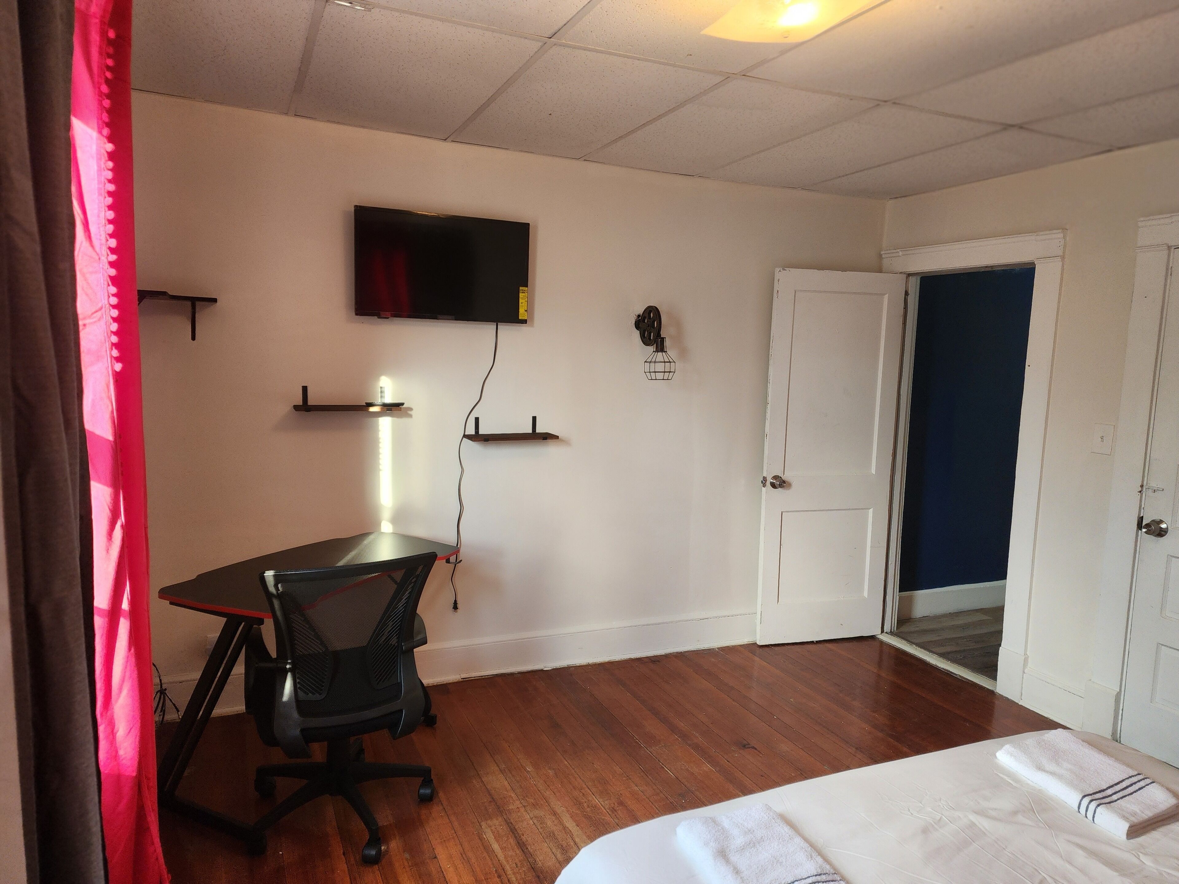 A room with a bed, a desk, a chair, a TV mounted on the wall, and a door leading to another room.