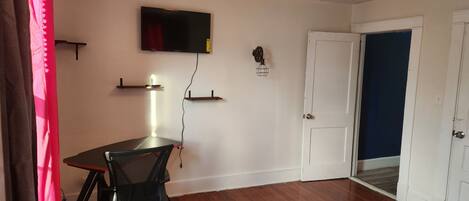 A room with a bed, a desk, a chair, a TV mounted on the wall, and a door leading to another room.