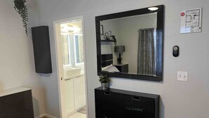 Ein Schlafzimmer mit einem Bett, einem Schminktisch, einem Spiegel, einer Pflanze und einer Tür zu einem weiteren Raum.